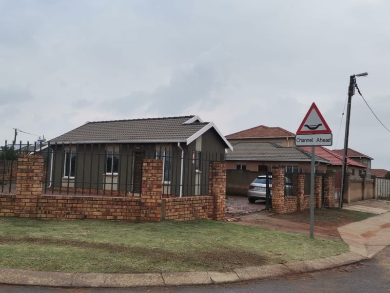 A front view of a single-story residential house featuring a brick perimeter wall, metal security fencing, and a paved driveway.