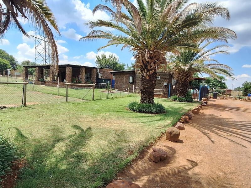 A view of the property exterior featuring brick buildings, a covered patio area, established palm trees, and a fenced garden space.