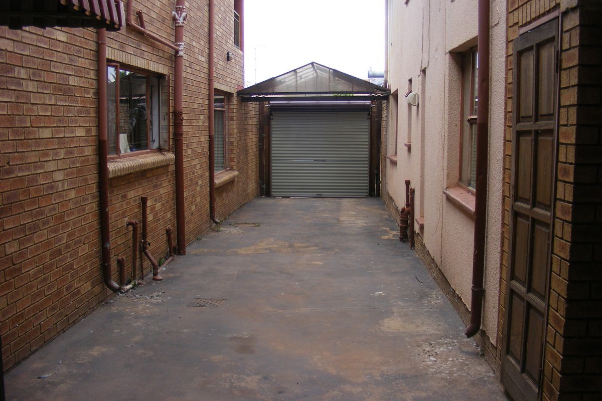 A narrow rear driveway area featuring a roller garage door, brick walls, and exposed downpipes, showing signs of wear and weathering.