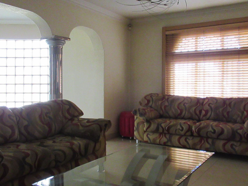 A living room featuring a distinctive glass block wall, decorative column, arched architectural details, and large windows with wooden blinds.