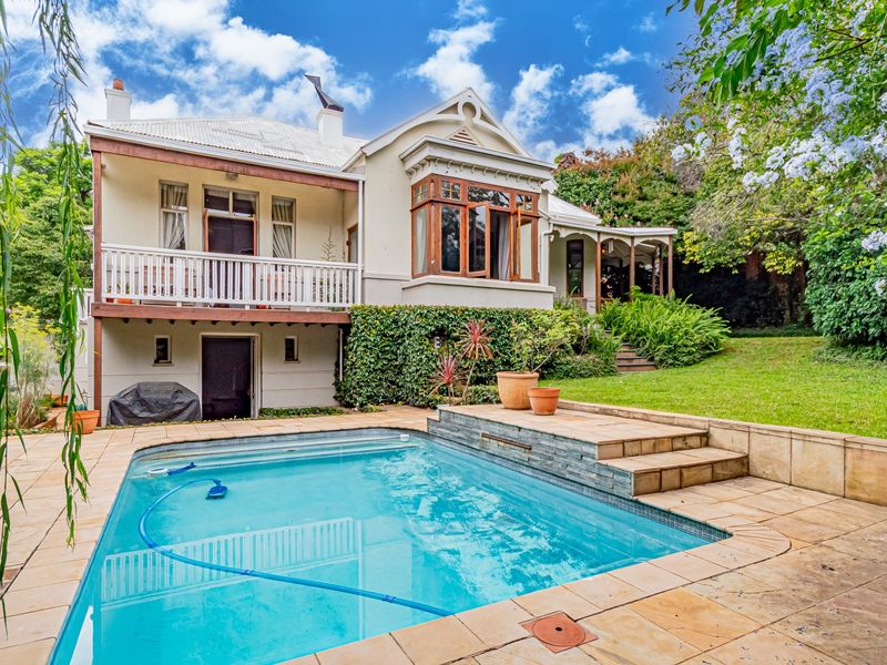 A well-maintained rear exterior view featuring a swimming pool with stone surround, a raised deck area, and a balcony overlooking the landscaped garden.