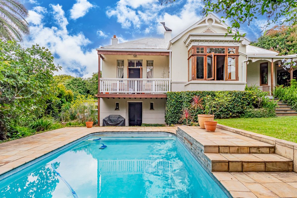 Rear view of a charming house featuring a swimming pool, stone-paved pool deck, and an elevated timber balcony overlooking the garden.