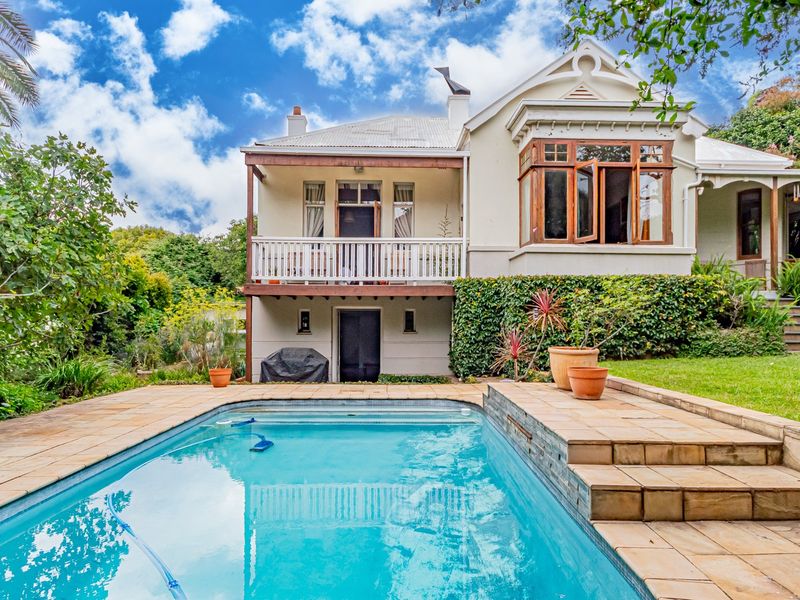 Rear view of a charming house featuring a swimming pool, stone-paved pool deck, and an elevated timber balcony overlooking the garden.