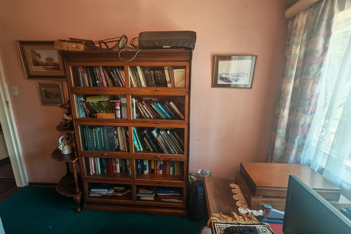 A bedroom space featuring fitted window security bars and curtain tracks, currently utilized as a home office area with dated wall finishes and carpeting.