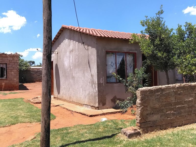 A modest single-story dwelling featuring a tiled roof and plastered exterior walls, situated on a plot with a brick boundary wall and grassed area.