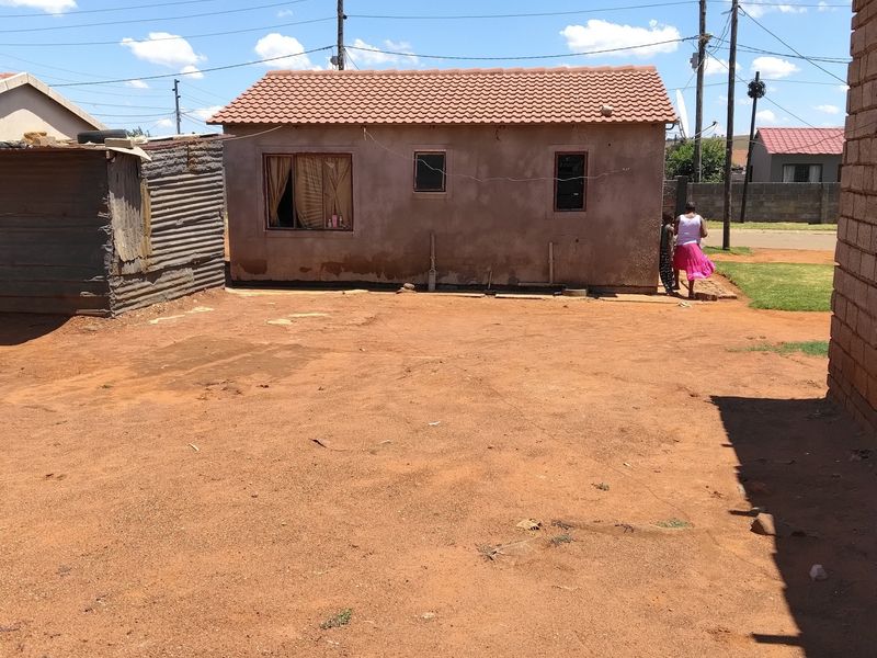Rear view of a small residential dwelling featuring a tiled roof and plastered masonry walls, set on an unpaved yard.
