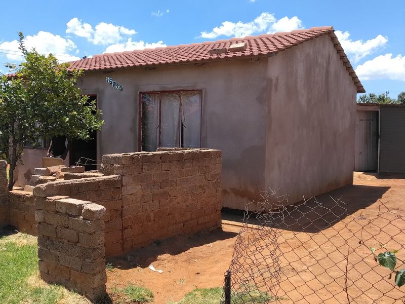Front view of a single-story house featuring a tiled roof, plastered exterior walls, and a partially enclosed yard with brick and wire fencing.