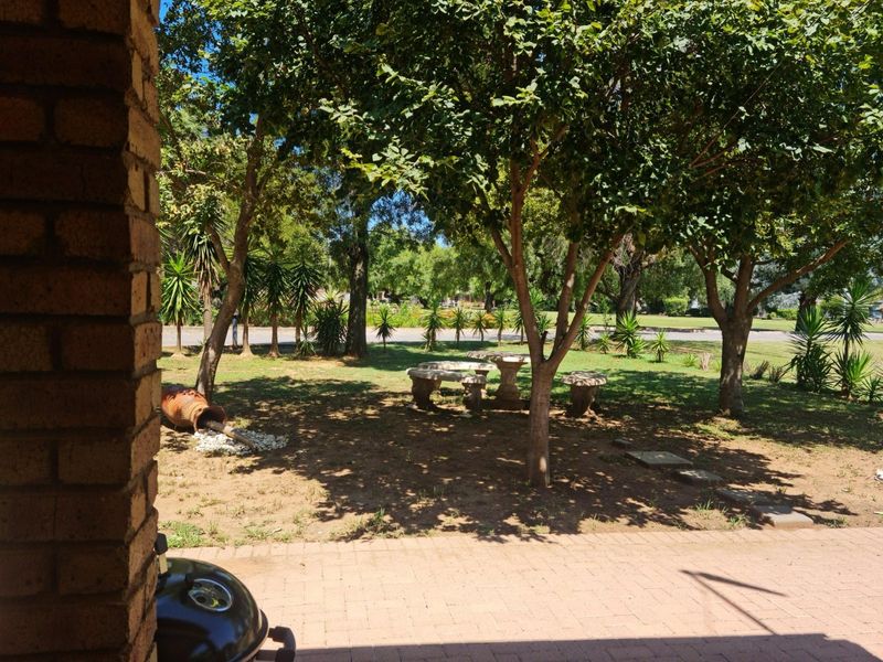 A spacious, tree-lined garden featuring established trees, a stone seating area, and brick-paved patio space, offering a peaceful outdoor setting.