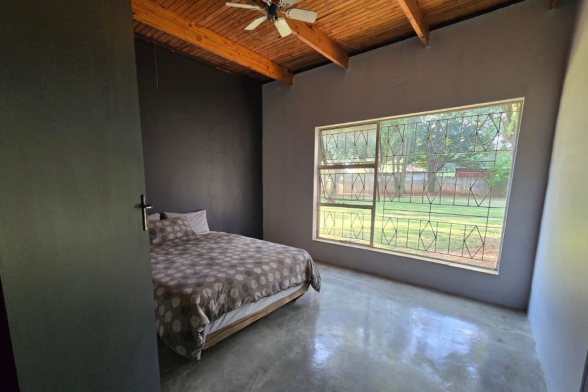A bedroom featuring polished concrete floors, exposed timber beam ceiling, and a large window with security bars overlooking the garden.