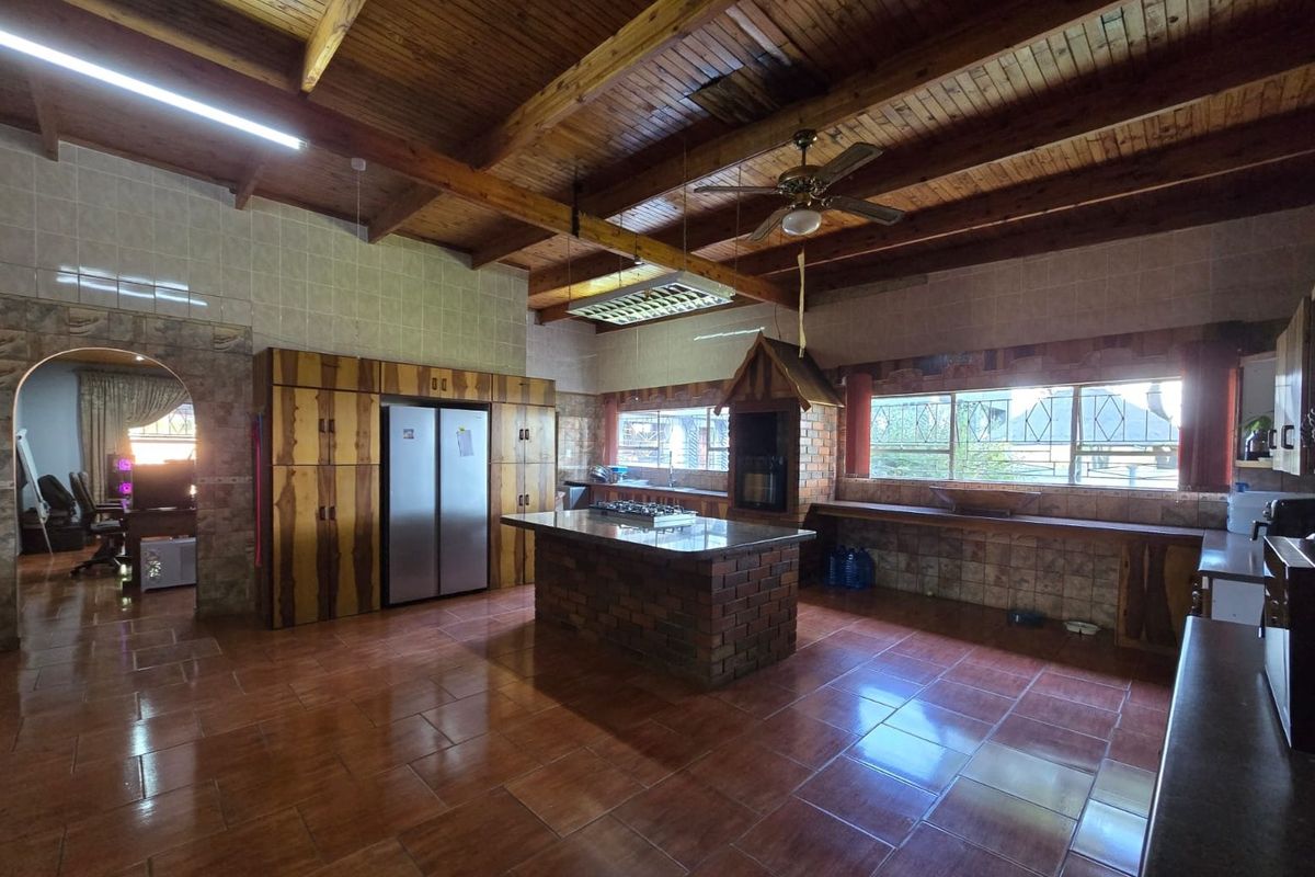 Spacious kitchen featuring exposed timber beam ceilings, a central brick island with built-in braai, and extensive tiled surfaces throughout.