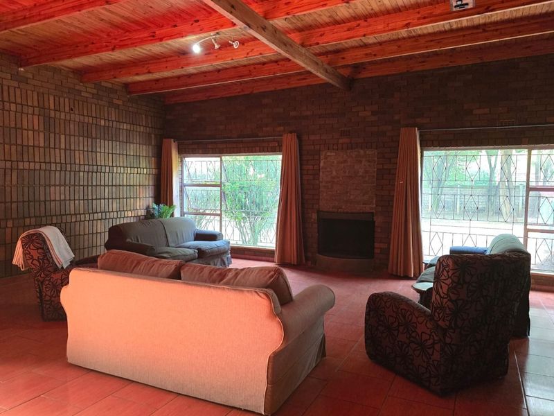 Spacious living room featuring rustic facebrick walls, a high exposed timber beam ceiling, tiled flooring, and a built-in fireplace with large windows.