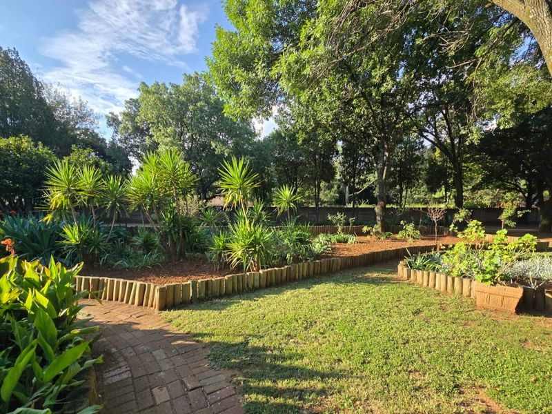 A well-maintained garden area featuring brick pathways, timber log edging, and established mature trees providing shade and greenery.