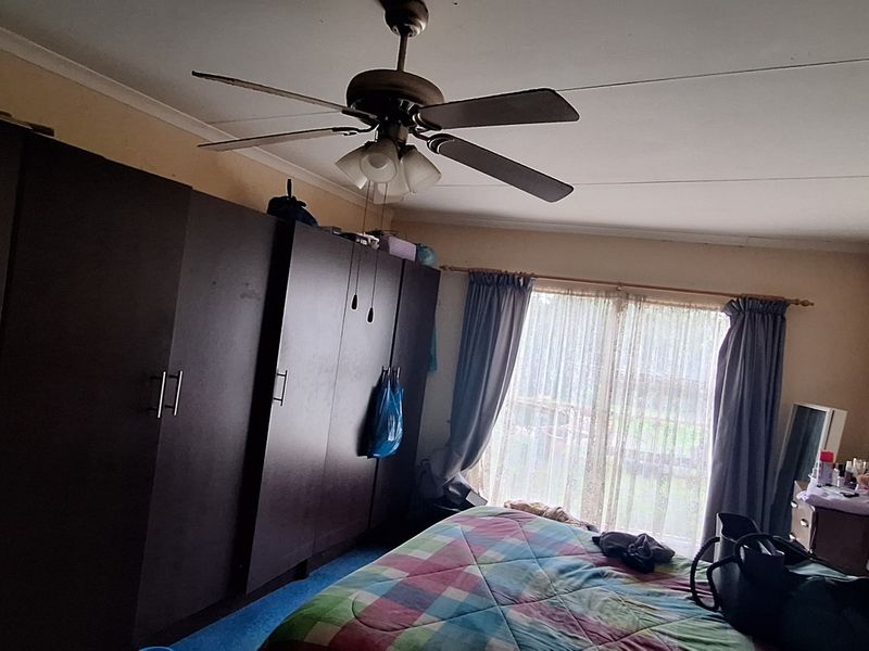 A bedroom featuring built-in cupboards, a ceiling fan, and large windows with curtain rails, requiring some cosmetic attention to the ceiling and walls.