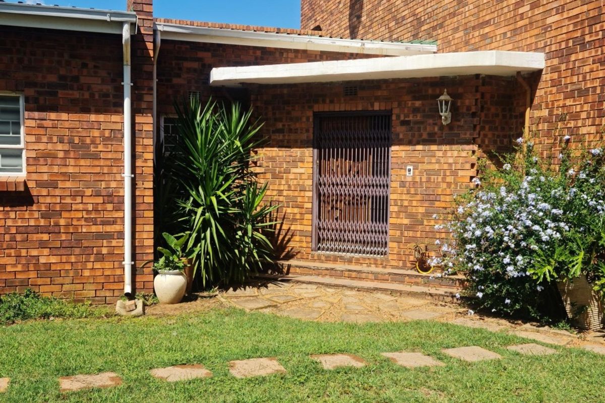 A rear exterior view of a brick house featuring a paved entrance area, security gate, and established garden surroundings.