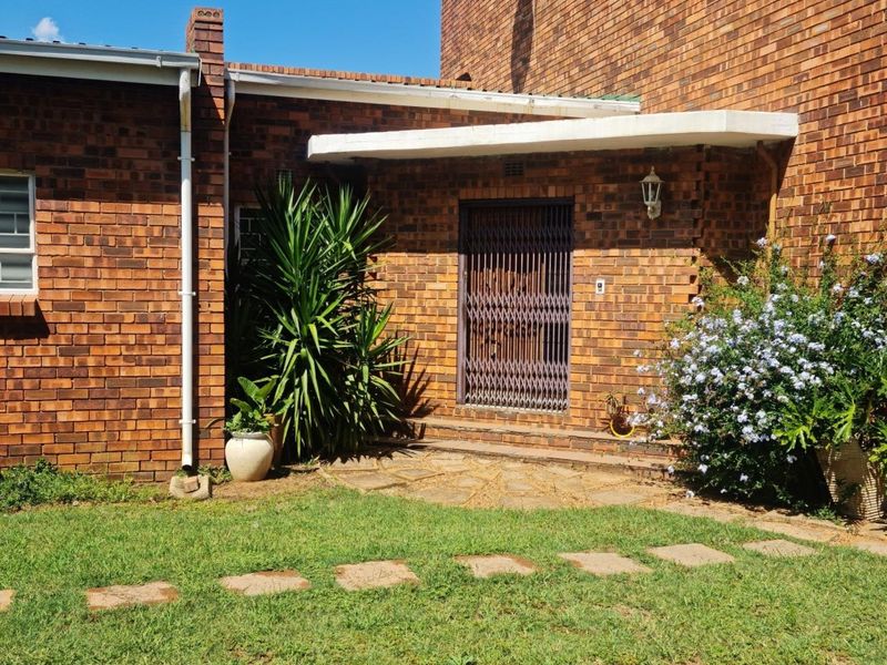 A rear exterior view of a brick house featuring a paved entrance area, security gate, and established garden surroundings.