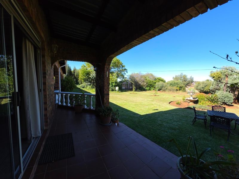 A covered brick patio featuring tiled flooring and a balustrade, overlooking a spacious, well-maintained garden with a lawn and fountain.