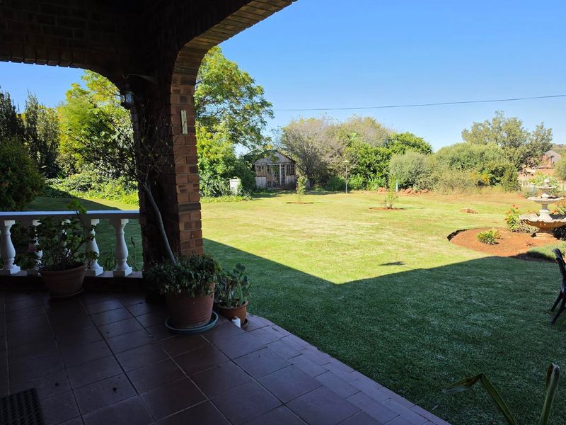 A covered brick patio overlooking a spacious, well-maintained garden with a water feature and mature trees.
