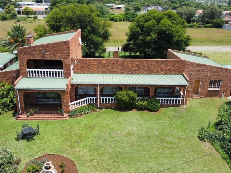 Aerial view of a face brick house featuring a green metal roof, covered patio, balcony with balustrades, and a spacious surrounding garden.