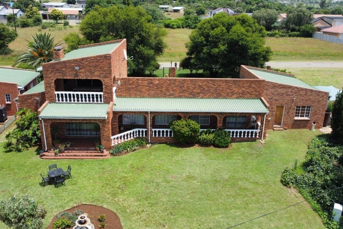 Aerial view of a face brick house featuring a green metal roof, covered patio, balcony with balustrades, and a spacious surrounding garden.