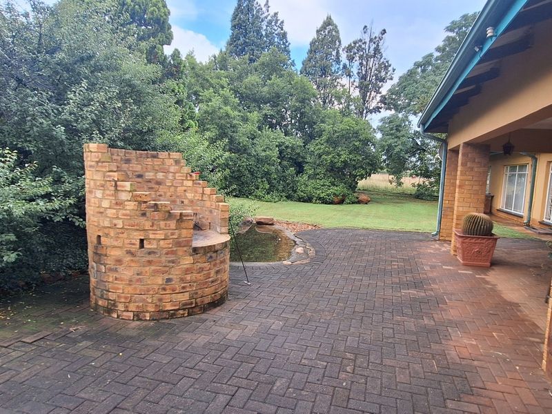 A spacious covered patio featuring a built-in brick braai, paved flooring, and a small water feature overlooking the garden.