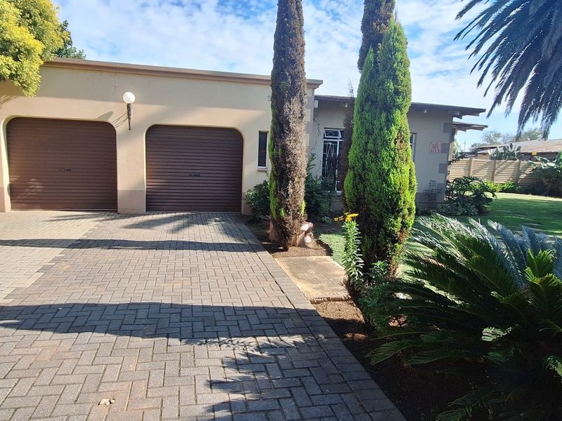 Front view of the property featuring a double garage, paved driveway, and established garden with mature trees and boundary walling.