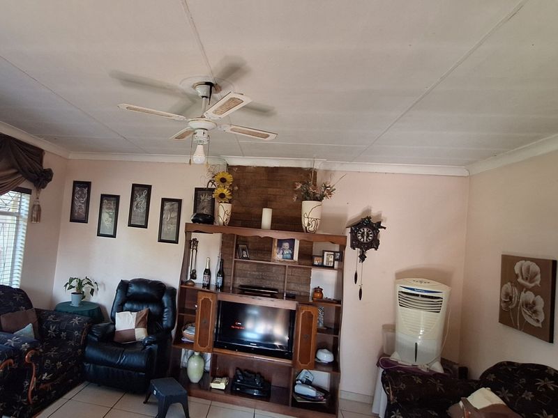 A living room featuring a brick accent wall, tiled flooring, and a ceiling fan, with visible wear on the ceiling panels.