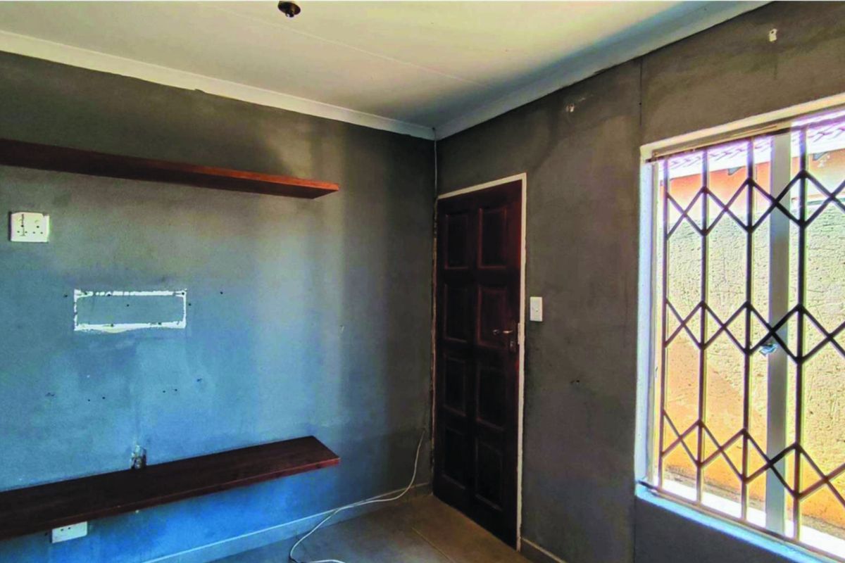 An empty living room featuring unfinished plastered walls, built-in wooden shelving, and a window fitted with a security gate.