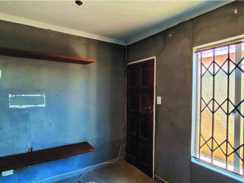 An empty living room featuring unfinished plastered walls, built-in wooden shelving, and a window fitted with a security gate.