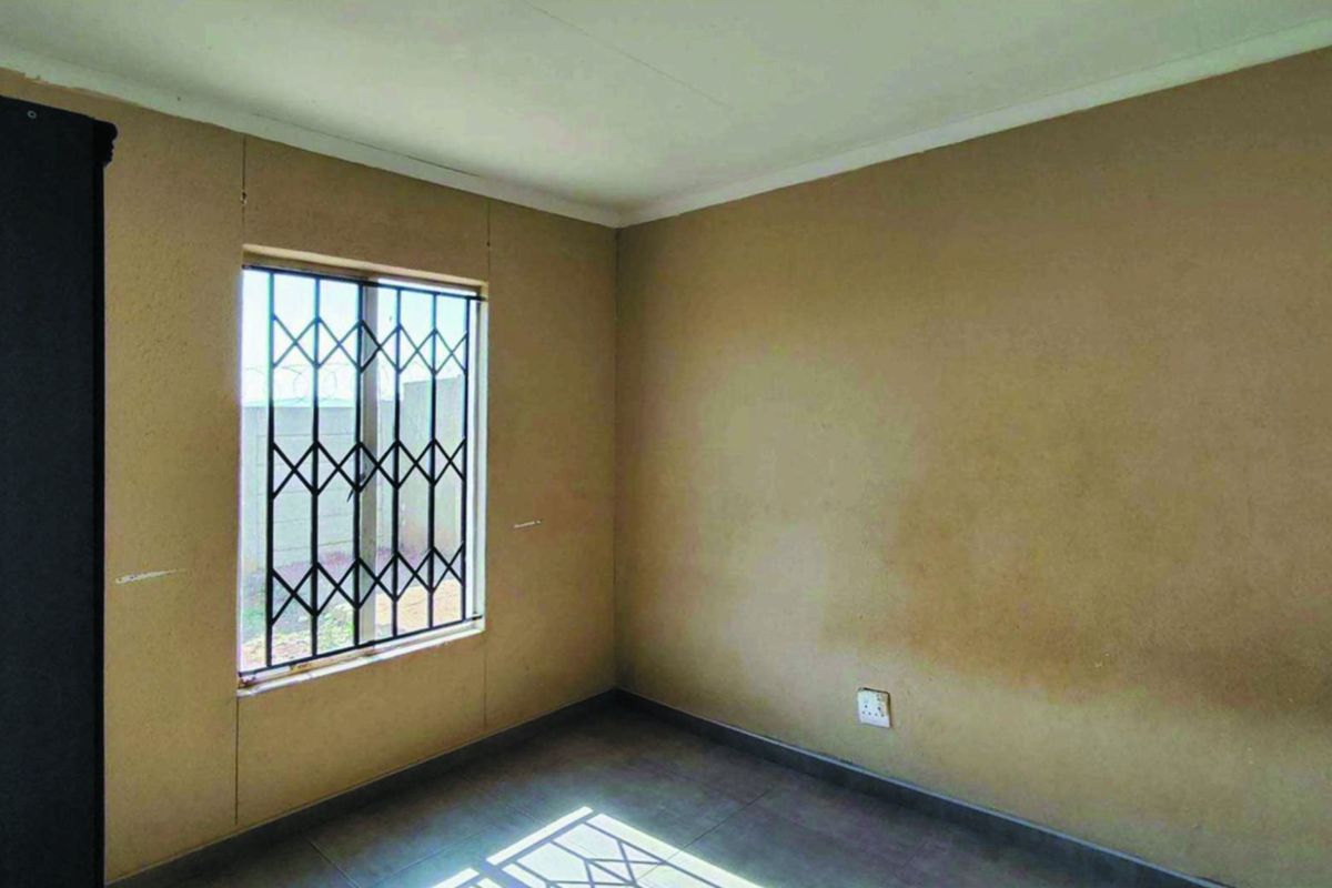 An empty bedroom featuring tiled flooring and a window fitted with a security gate, requiring cosmetic updates to the wall finishes.