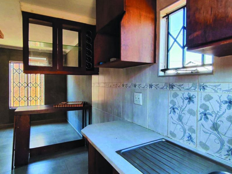 A dated kitchen featuring built-in wooden cabinetry, a stainless steel sink, and decorative tiled backsplash with security bars on the windows.