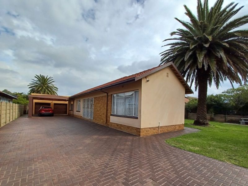 A well-maintained single-story house featuring a paved driveway, attached carport, and established garden with a large palm tree.