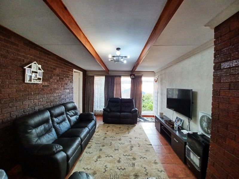 A living room featuring exposed timber ceiling beams, a rustic brick feature wall, and tiled flooring with direct access to the outdoor area.