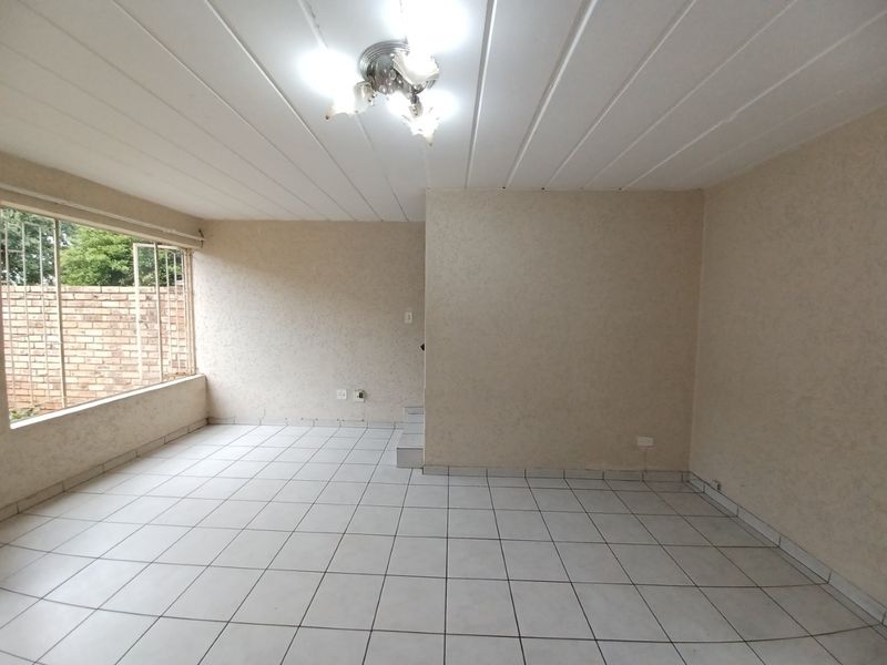 An empty, open-plan living area featuring tiled flooring, a panelled ceiling, and a large window with security bars.