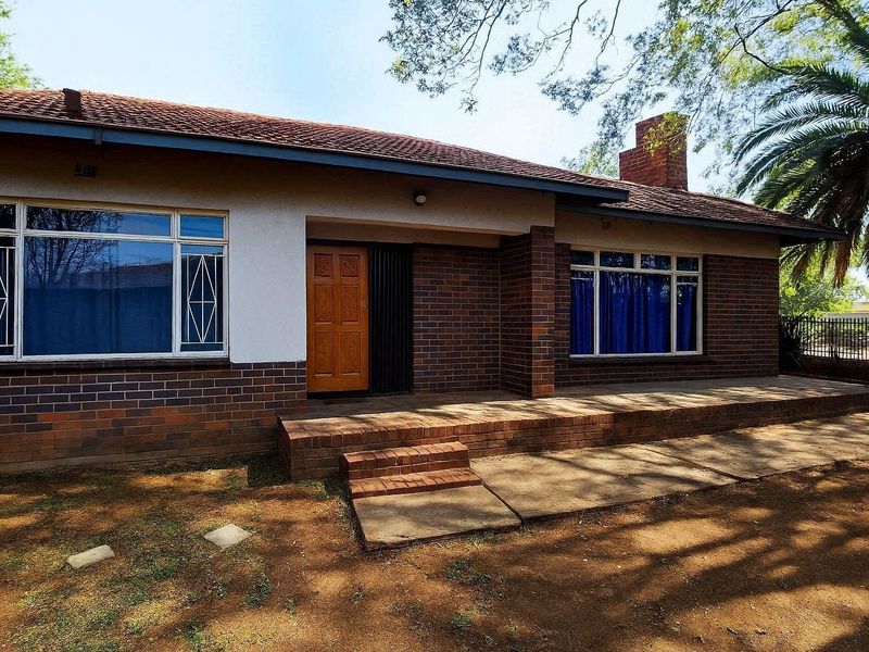 Front elevation of a single-story brick house featuring a covered porch, tiled roof, and established garden surroundings.