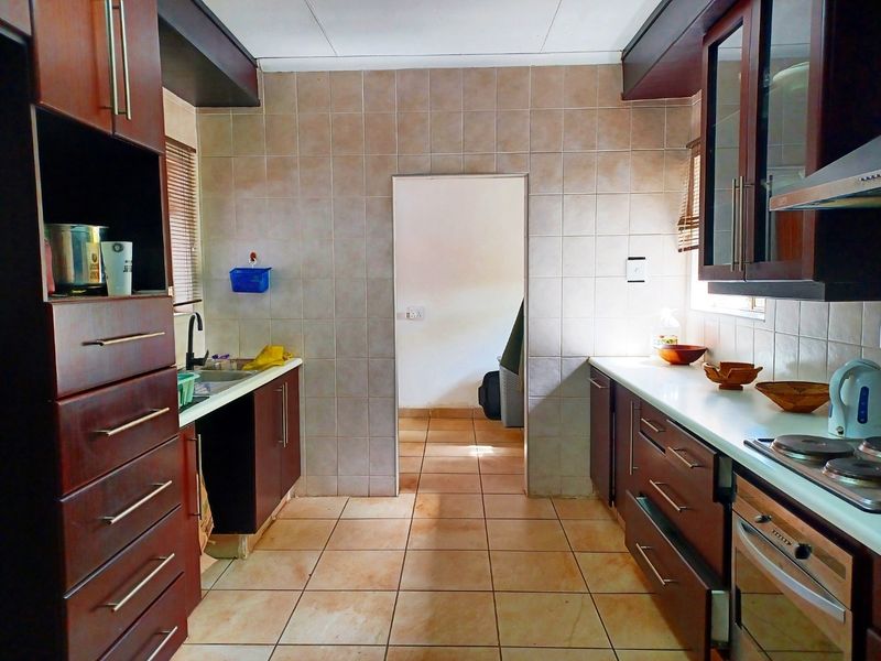 A functional kitchen featuring dark wood cabinetry, tiled splashbacks, built-in oven, electric hob, and tiled flooring with a central doorway leading to an adjacent room.