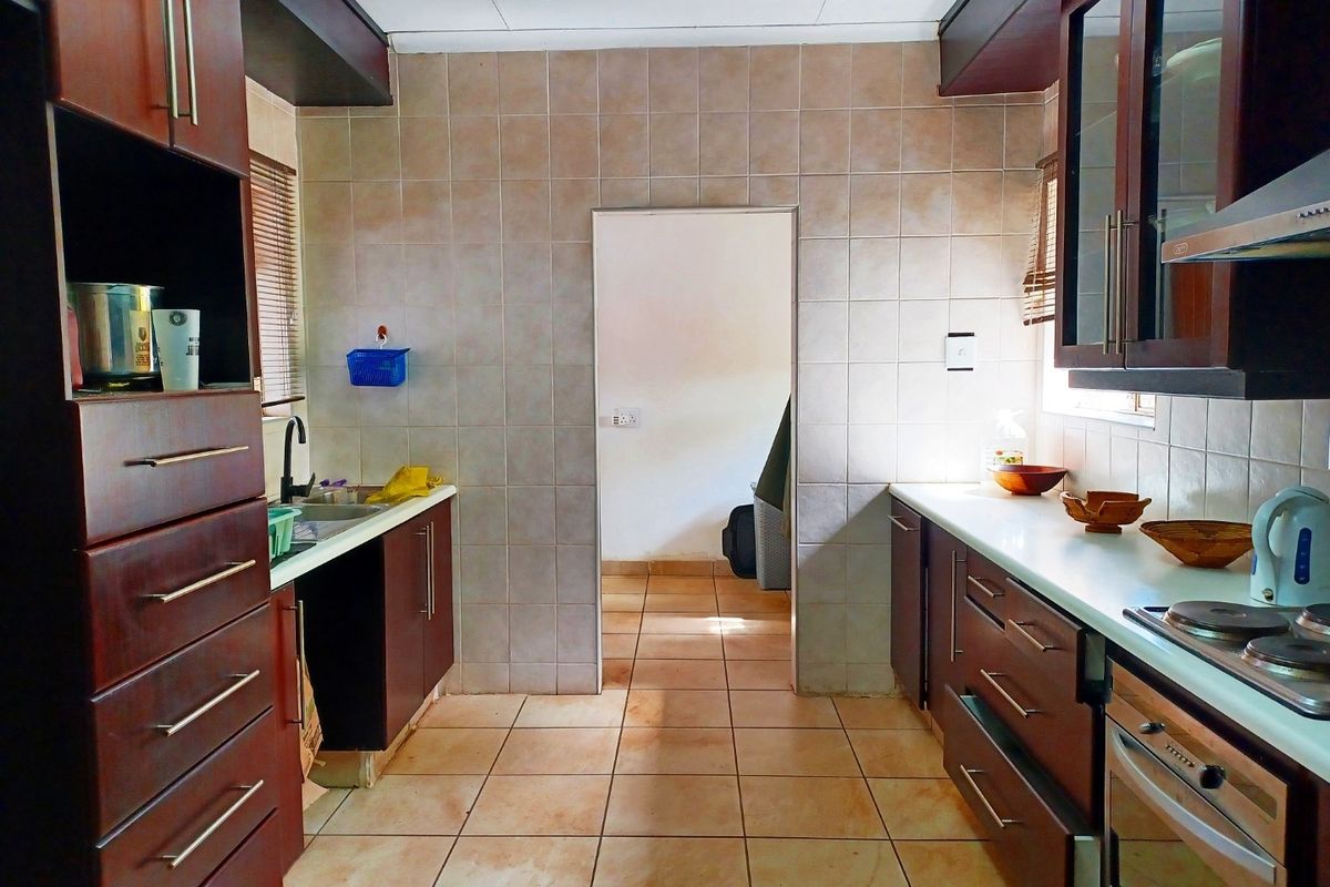 A functional kitchen featuring dark wood cabinetry, tiled splashbacks, built-in oven, electric hob, and tiled flooring with a central doorway leading to an adjacent room.