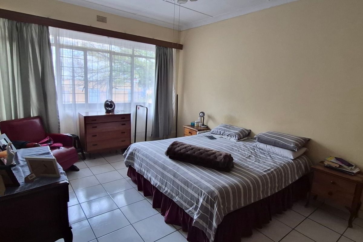 A spacious bedroom featuring tiled flooring, a large window with curtain rails, and a ceiling fan, offering a functional and well-lit living space.