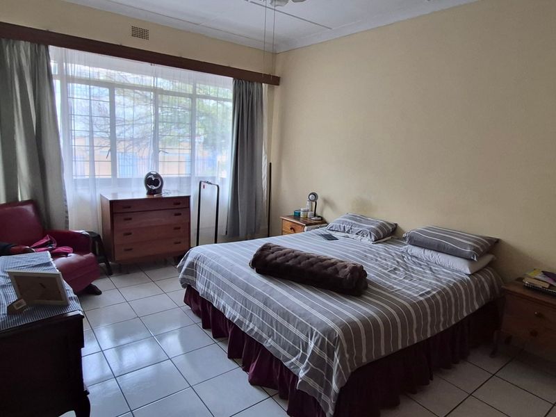 A spacious bedroom featuring tiled flooring, a large window with curtain rails, and a ceiling fan, offering a functional and well-lit living space.