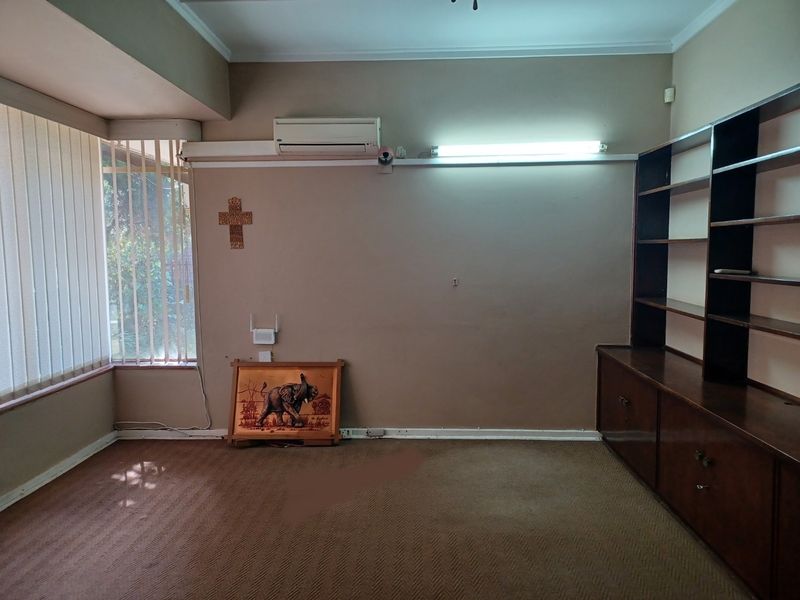 A spacious living area featuring built-in wooden shelving, a split-system air conditioner, and large windows with vertical blinds.