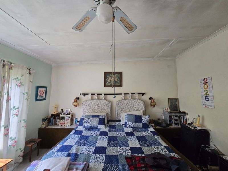 A bedroom featuring a built-in headboard, wall-mounted lighting, and a ceiling fan. The room shows signs of wear, including visible ceiling panel cracks.