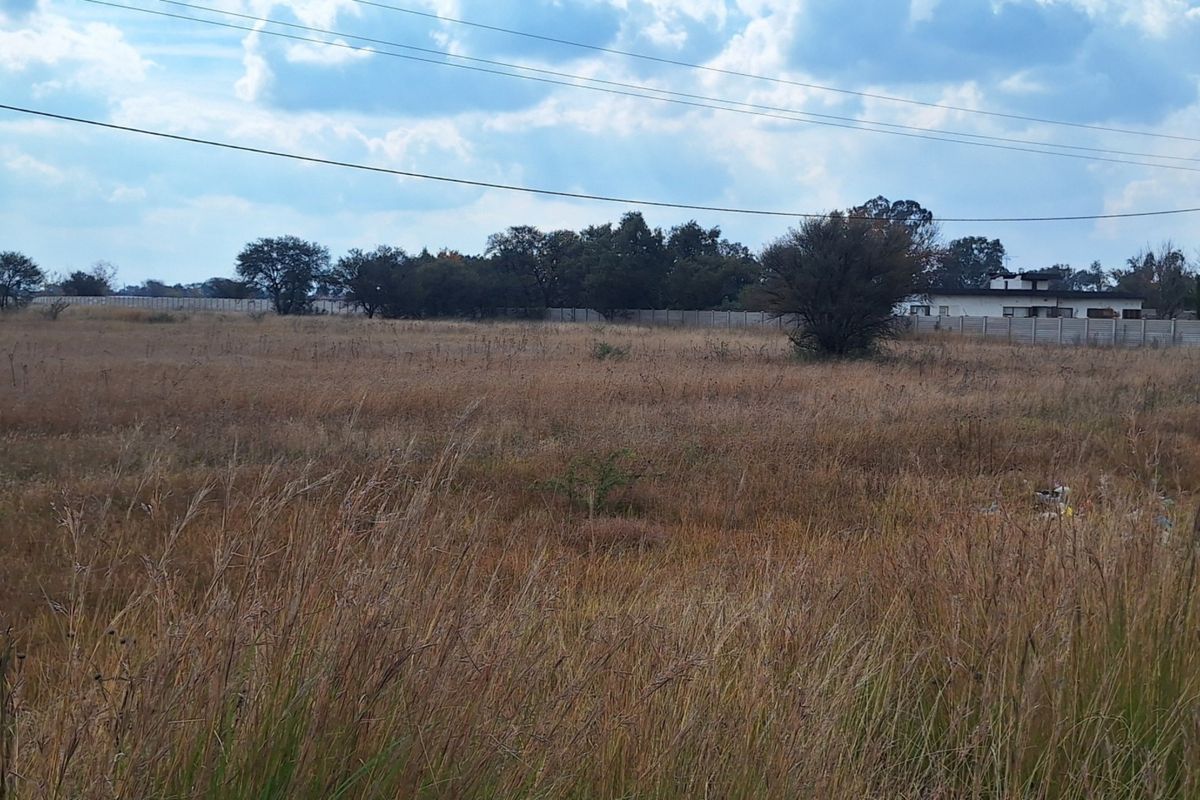 A wide-angle view of a vacant residential plot featuring dry grass, perimeter walling, and overhead power lines.