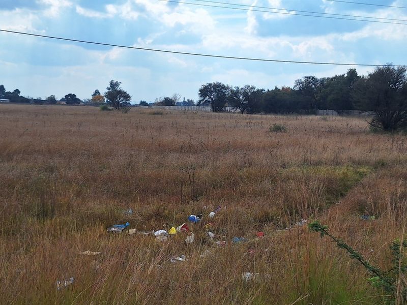 A view of a vacant residential plot featuring dry grass, scattered debris, and overhead power lines.