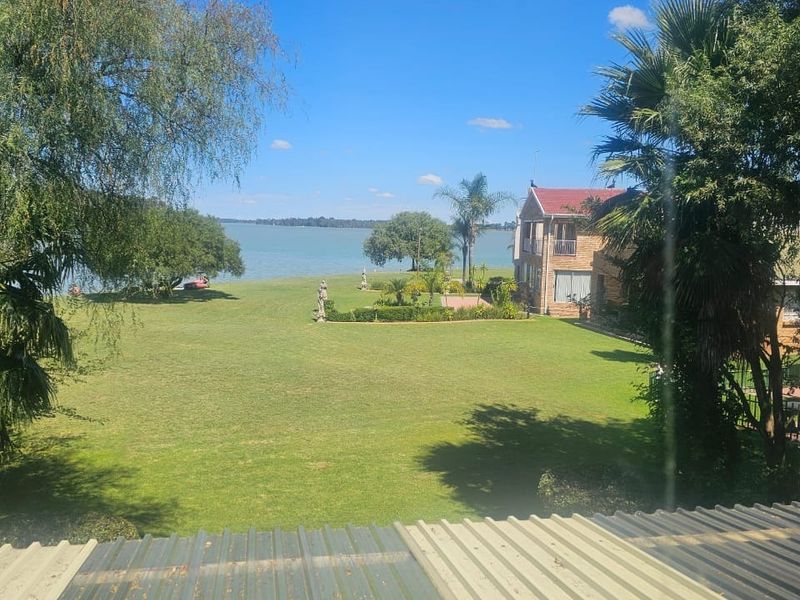 A scenic view overlooking a large green lawn leading down to the waterfront, framed by mature trees and a neighboring property.