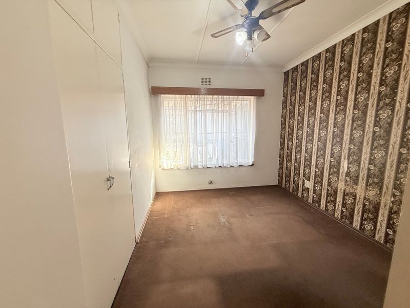 An empty bedroom featuring built-in cupboards, a ceiling fan, and a window with security bars, requiring cosmetic updates to the flooring and wall finishes.