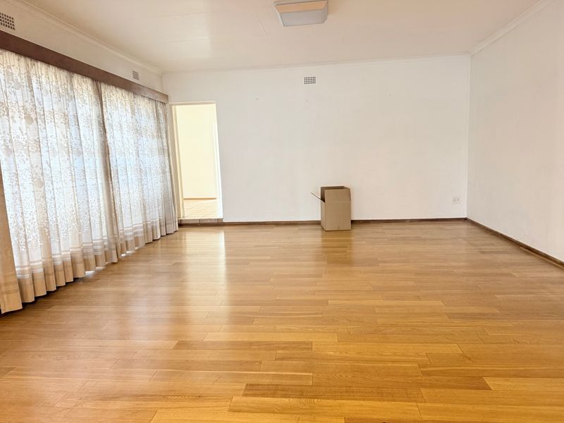 A spacious, empty living room featuring light-toned laminate flooring, neutral walls, and large windows with installed curtain tracks.