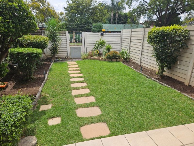 A well-maintained rear garden featuring a manicured lawn, stone paver pathway, and secure precast concrete boundary walls with a metal pedestrian gate.