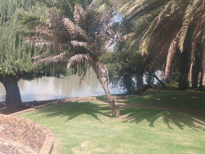 A scenic garden area featuring a curved brick retaining wall, mature trees, and direct access to the riverfront.
