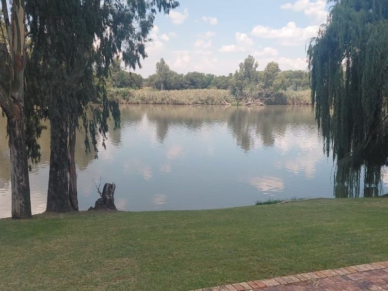 A scenic view of the riverfront featuring mature trees, a well-maintained lawn, and brick paving along the water's edge.
