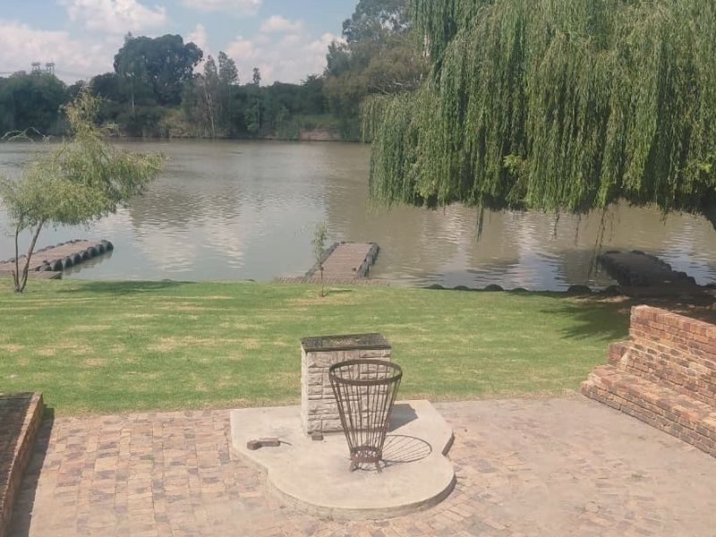 A scenic garden area featuring a brick-paved patio with a built-in braai, overlooking a river with private floating jetties.