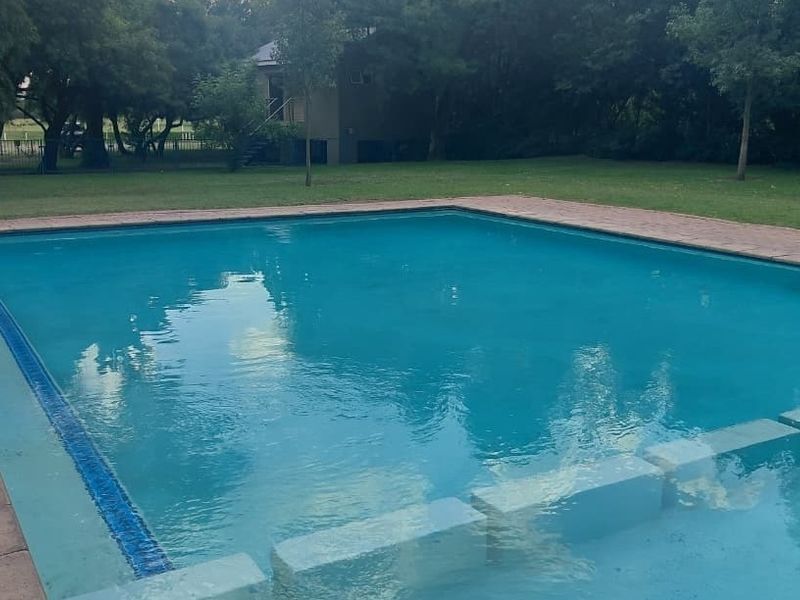 A well-maintained swimming pool set within a spacious garden area, featuring paved surrounds and established trees in the background.